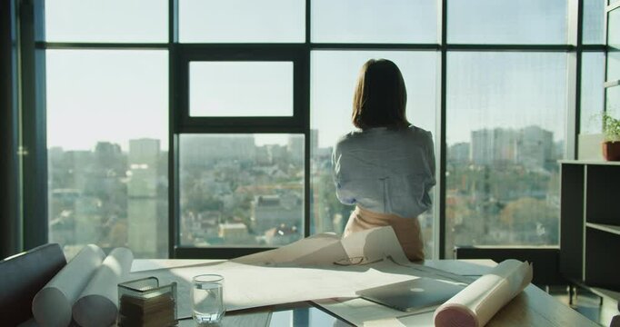 An architect standing by a window overlooking a cityscape, reflecting on project plans with blueprints and tools spread on the desk in a modern workspace
