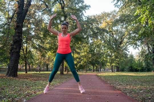 A mature, fit woman and man doing jumping jacks in a city park during summer, enjoying an active lifestyle with energy, fitness, and physical exercise outdoors.	