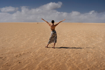 Obraz premium Joyful woman stands in sandy dessert of Fuerteventura, arms raised in pure happiness. clouds, sun, and her all-inclusive wristband symbolize ultimate freedom, relaxation, and perfect holiday escape