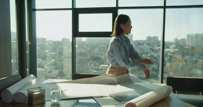 A professional architect reviewing project blueprints on a table with a laptop in a modern office, large windows framing an urban cityscape in the background