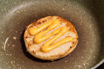 A sesame seed bun with zigzag mustard lines rests on a skillet, surrounded by crumbs and sesame...