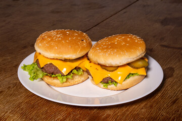 Two cheeseburgers with beef patties, melted cheese, lettuce, and pickles on sesame buns, served on a white plate atop a rustic wooden surface.