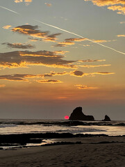 sunset over the sea with a red sun by a shark tooth looking rock with clouds and a trail left behind from a plane
