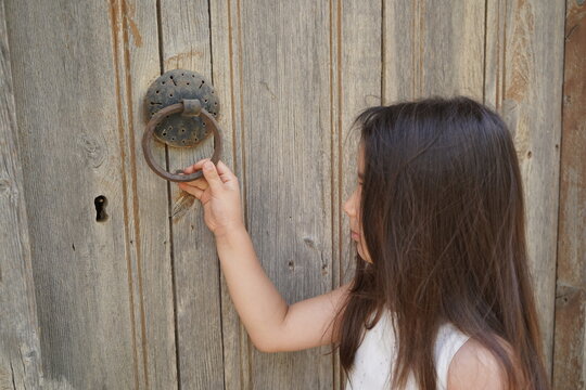 A five year old girl with brown long hair in front of an old wooden door holds the vintage handle ready to knock - Abandoned house in Raftis village in Crete, Greece