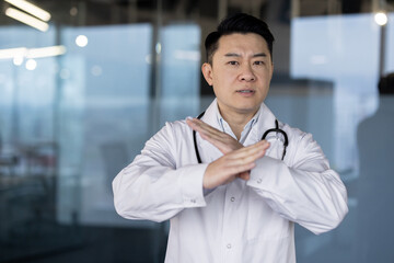 Fototapeta premium Portrait of a serious young and worried Asian male doctor in a white coat standing in the clinic lobby and showing a no stop sign with his hands at the camera