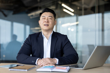 Relaxed young Asian male businessman sitting at the desk in the office, eyes closed, breathing deeply and resting