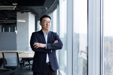 Fototapeta premium Asian young male businessman standing in the office near the window wearing a suit and glasses, arms crossed on his chest and looking to the side