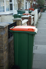 A green trash bin with an orange lid for recyclable waste stands on the street in the city, with buildings and other trash bins in the background.