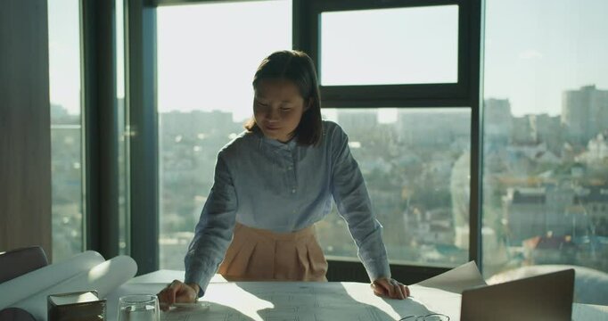 A dedicated architect reviewing and sketching blueprints on a desk in a modern, sunlit office with panoramic city views, representing creativity and professionalism