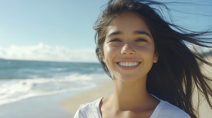 Portrait of young woman at sea looking at camera. Smiling latin hispanic girl standing at the beach with copy space and looking at camera. Happy mixed race girl in casual outfit with wind in her hair