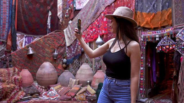 Tourist cowgirl vlogging with phone in authentic shop environment consisting of colorful carpets and rugs hanging from the wall.