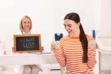 Happy woman lost weight and nutritionist with small blackboard in clinic, selective focus