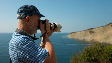 Obraz premium Adult gray-haired man with a camera at the eye on the background of a sea bay. The camera has a long lens with a hood.