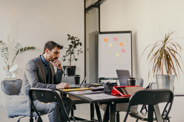 The businessman is wearing a suit, working at his desk, and using a laptop in the office.