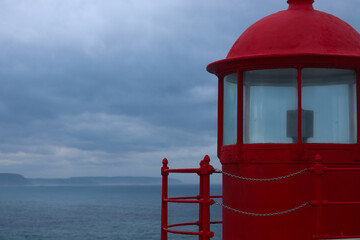 A red lighthouse at Forte de Sao Miguel Arcanjo in Nazare, Portugal. The historic beacon stands against a cloudy sky, overlooking the Atlantic Ocean, guiding sailors near the famous big wave surf spot