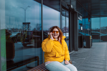 Fototapeta premium Happy stylish young 30s woman wearing trendy outfit talking on cell phone, making call and sitting outdoors on modern city street. Urban lifestyle concept. High quality photo