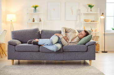 Unwell man lying on the sofa at home living room, experiencing stomach pain, cramps, discomfort, or constipation. Challenges of dealing with health issues and the need for relief hurt.