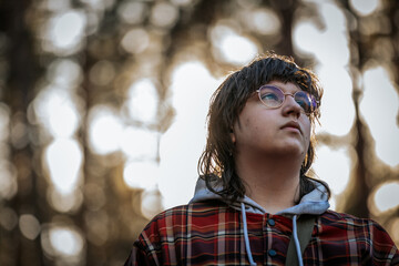 A young person wearing glasses, a plaid jacket, and a hoodie gazes upward with a contemplative...