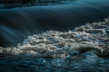 A powerful wave crashes in a river, with white foam and dark blue water creating a dramatic contrast. The movement and textures highlight the dynamic energy of flowing water.