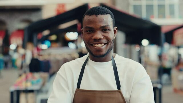 Portrait of smiling African American service worker in apron crossing arms while looking at camera standing near restaurant