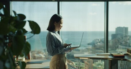 Young professional woman using a laptop in a bright office with a panoramic ocean view, working while standing near large windows in a modern workspace