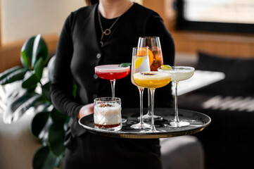 A bartender presents a variety of colorful cocktails on a black tray. The drinks include a refreshing mix of flavors, garnished with fruits, perfect for a lively gathering.
