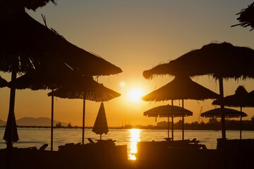 Scenic beach sunset with silhouetted umbrellas and calm sea. Golden sunlight reflects on the water, creating a relaxing atmosphere with boats and a distant harbor view.