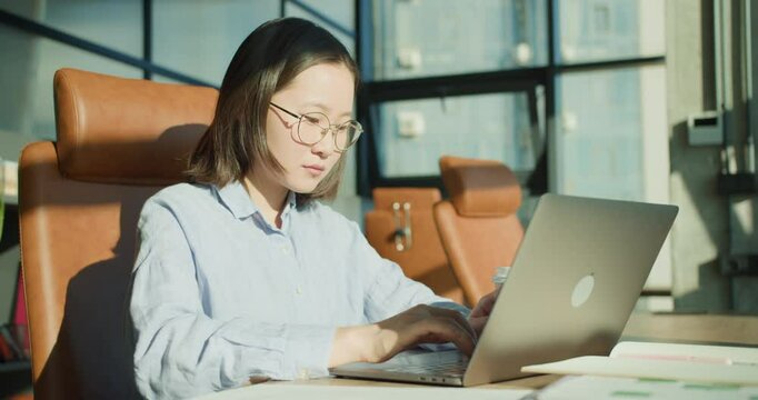 A focused young woman working on her laptop in a modern office setting with sunlight highlighting her determination and creating a productive atmosphere
