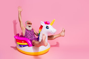 Joyful senior man with beard wearing funky leopard print bodysuit, having fun on inflatable unicorn against a vibrant pink backdrop