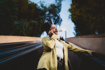 young Hispanic woman smiles while talking on phone as she rides an outdoor escalator at dusk. Dressed in green shirt, female looks content and engaged in conversation, with city lights in background