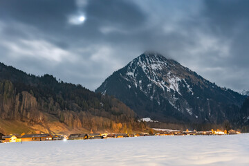 Winter night landscape in the mountains, full moon hidden behind clouds over the mountains. Small village in the Alps, houses lit at night, view from a hiking trail.at night, view from a hiking trail.