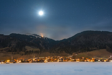 Winter night landscape in the mountains, full moon in the starry sky above the mountains. Small village in the Alps, houses illuminated at night, view from a hiking trail.