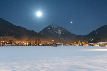 Winter night landscape in the mountains, full moon and planet Venus in the starry sky above the mountains. Small village in the Alps, houses illuminated at night, view from a hiking trail.
