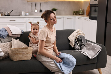 Tired housewife with her little daughter among messy laundry on sofa at home