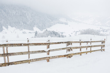 Snow-covered wooden fence on a mountain pasture. Winter landscape on a mountain hiking trail on a cloudy and foggy day.