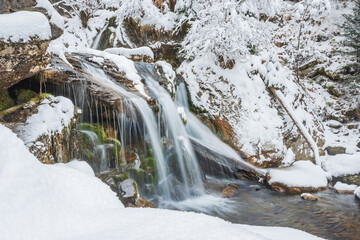 Small waterfall on a mountain river, water flows down snow-covered rocks. Winter landscape on a mountain hiking trail.