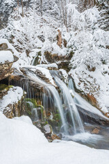 Small waterfall on a mountain river, water flows down snow-covered rocks. Winter landscape on a mountain hiking trail.