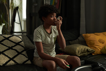 Cute youthful boy pumping inhaler in his mouth during asthma attack while sitting on couch and...