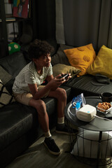 African American boy with joystick in hands sitting on couch in front of tv set in living room and playing video game
