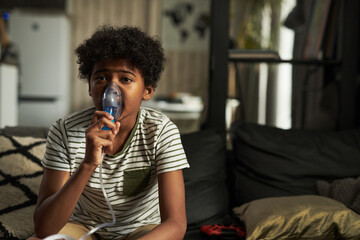 Adolescent boy in t-shirt watching tv broadcast and inhaling fumes from electric nebulizer while spending time at home