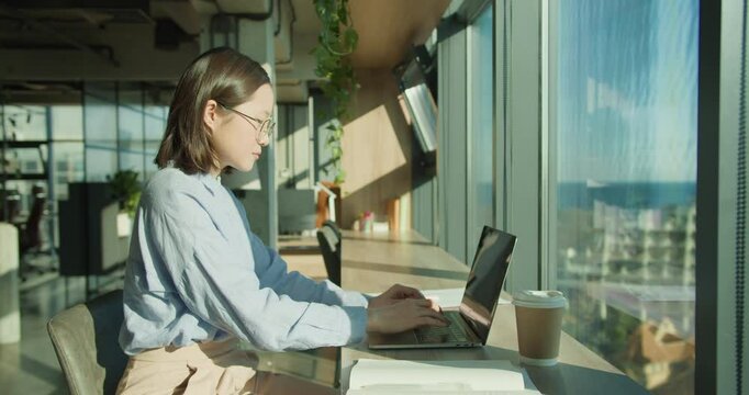 A young professional woman working on a laptop in a sunlit office with modern decor, surrounded by plants and enjoying a coffee, showcasing productivity and inspiration
