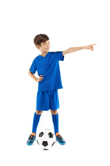 Cheerful young boy in blue sport outfit posing with classic soccer ball pointing finger away over white background