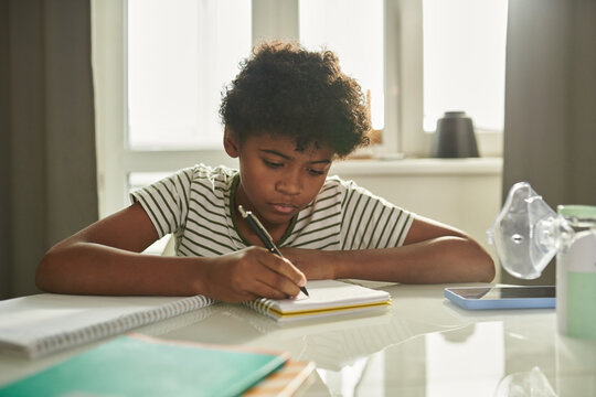 Youthful serious schoolboy bending over copybook or notepad and taking notes while carrying out assignment in home environment
