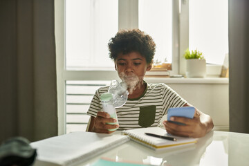 Asthmatic schoolboy with inhaler breathing vapors from mask while sitting by table at home and carrying out schoolwork