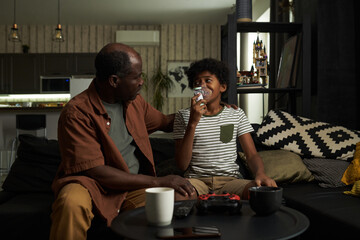 Senior African American man looking at youthful grandson while helping him with inhaler during asthma attack