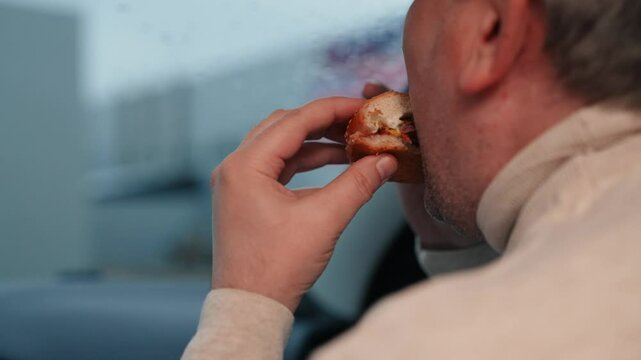 Man takes bite of tasty burger to quell hunger sitting in car on cloudy day. Man consuming burger in car and enjoying view from window. Man indulging in unhealthy burger in car in move on road of city