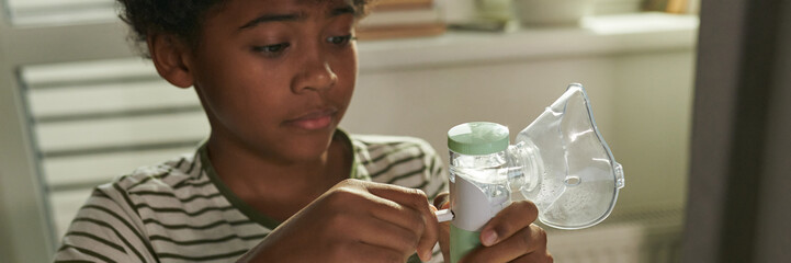 Header of schoolboy with asthma plugging inhaler with oxygen mask while preparing for medical treatment at home