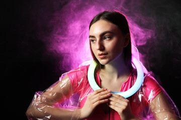 Naklejka premium Beautiful woman with ring lamp wearing transparent coat on dark purple background in smoke