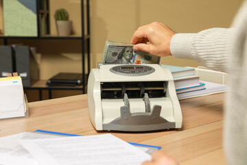 Man putting money into banknote counter at wooden table indoors, closeup