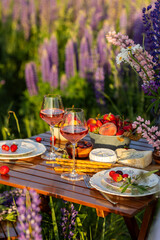 Romantic table decor for a loving couple on blooming meadow with purple lupines. Two glasses of wine, flowers in a vase, silverware, fruits, wooden furniture. Sunset, golden hour, atmospheric light